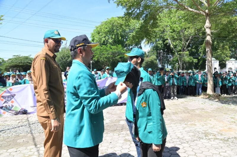 Staf Ahli Bupati Aceh Besar, Fata Muhammad SPd MM memasang topi kepada Mahasiswa KKN USK kepada di halaman Dekranasda, Gampong Gani, Kecamatan Ingin Jaya, Aceh Besar, Selasa (07/07/2026). FOTO/MC ACEH BESAR